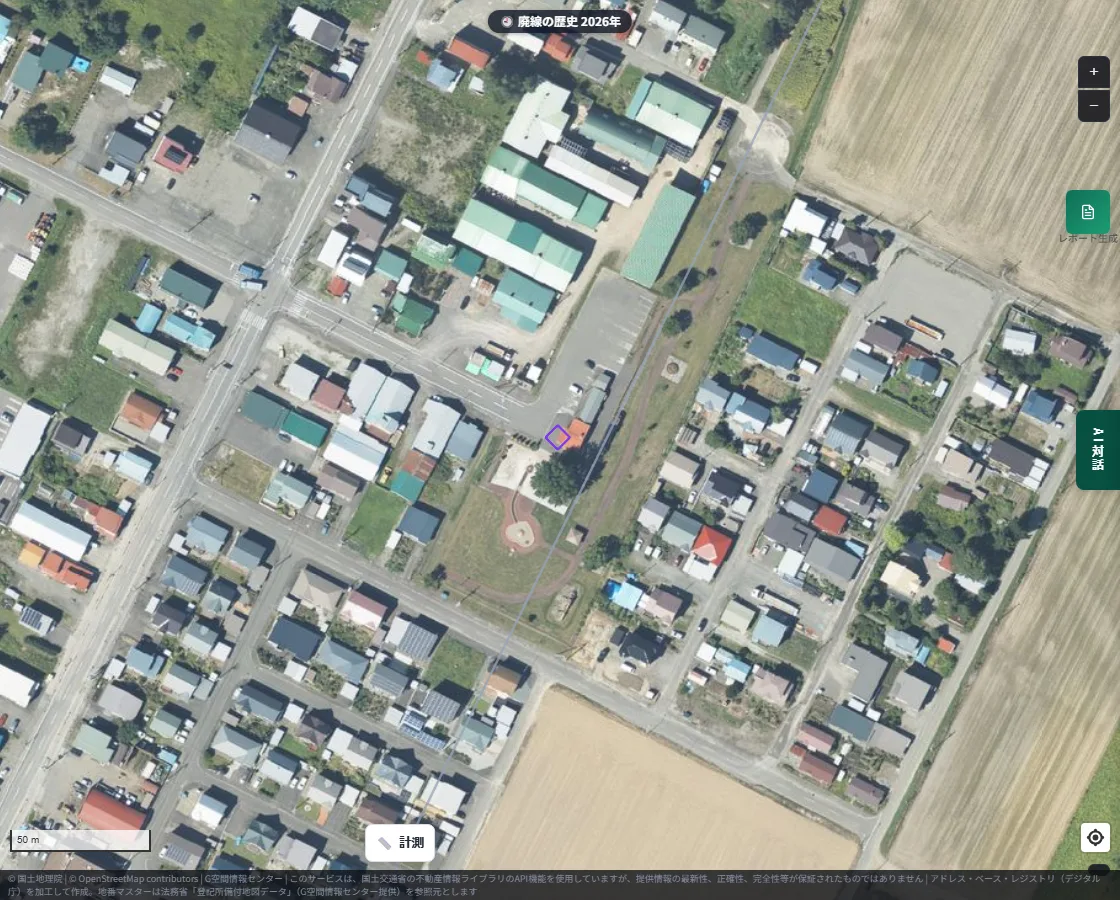 A zoomed-in aerial photograph of the former Aikoku Station site. Around the former-station position marked by a purple frame, a park-like site with curved paths is visible, surrounded by houses and warehouses