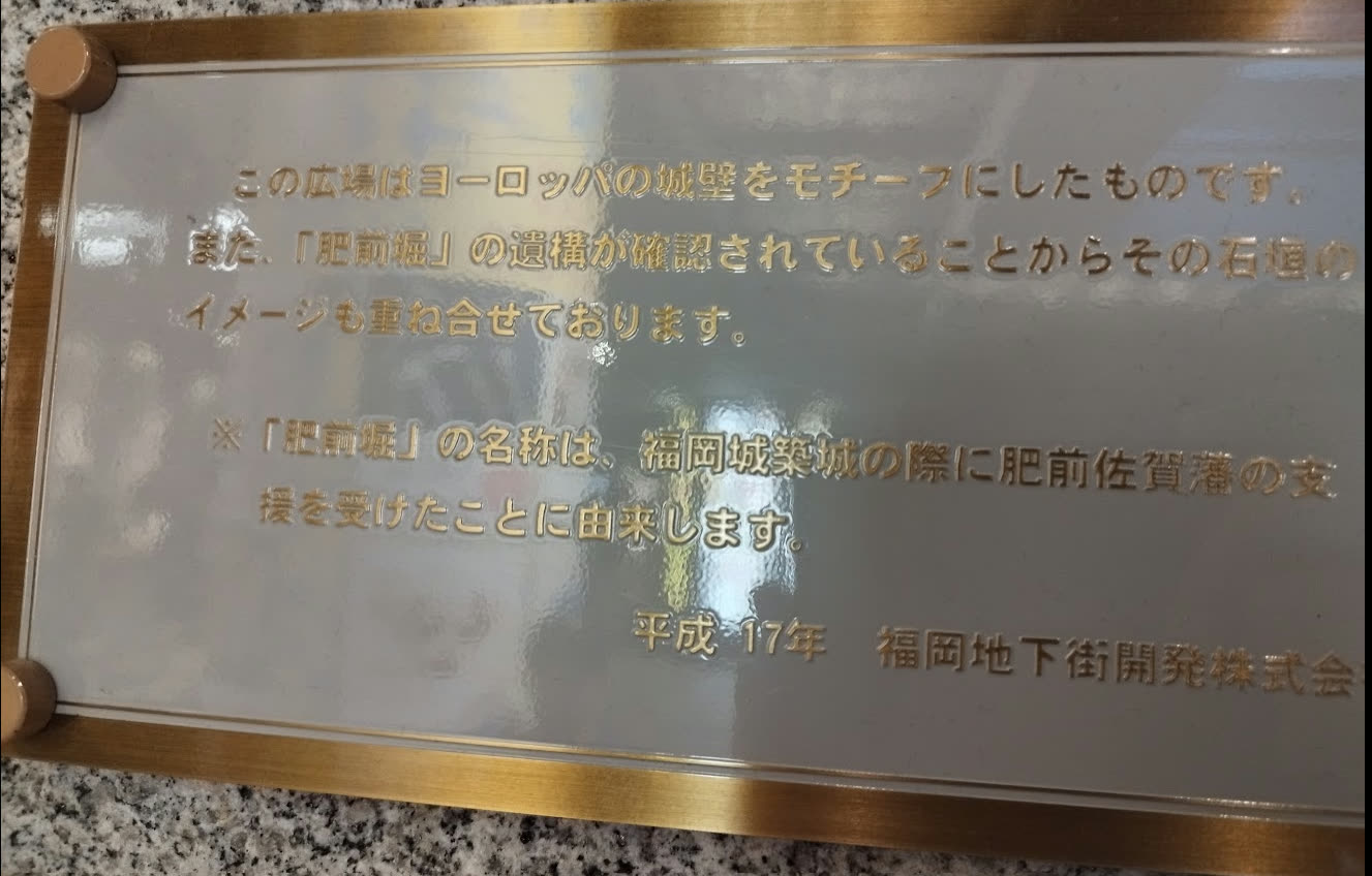 Plaque for the "Stone-Paved Plaza," explaining that the plaza is modeled on European castle walls, that it also layers in the image of stonework from the confirmed remains of the "Hizen Moat," and describing the origin of the name "Hizen Moat"