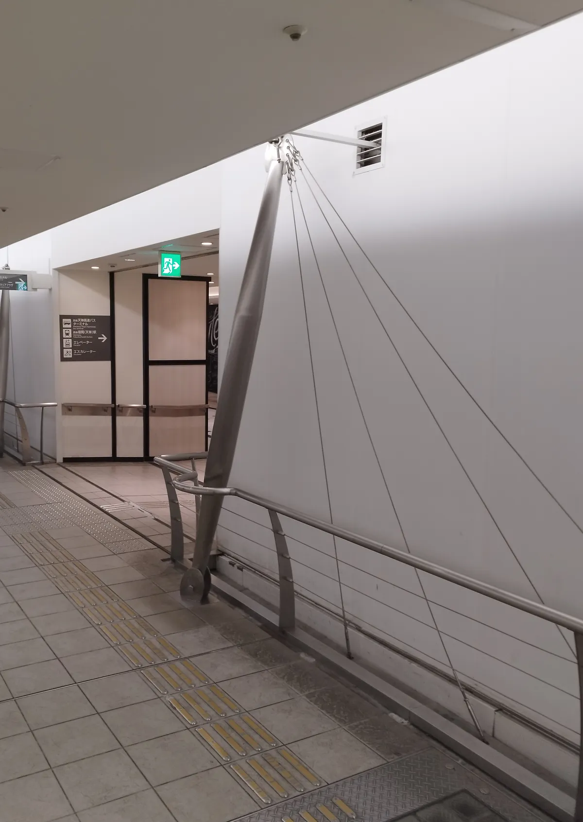 A slanted metal pylon with cables fanning out across the white wall, and a bridge-like handrail running in the foreground